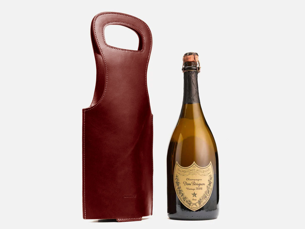 A bottle of Dom Pérignon champagne stands next to a tall, brown leather wine carrier with a handle, set against a plain white background.